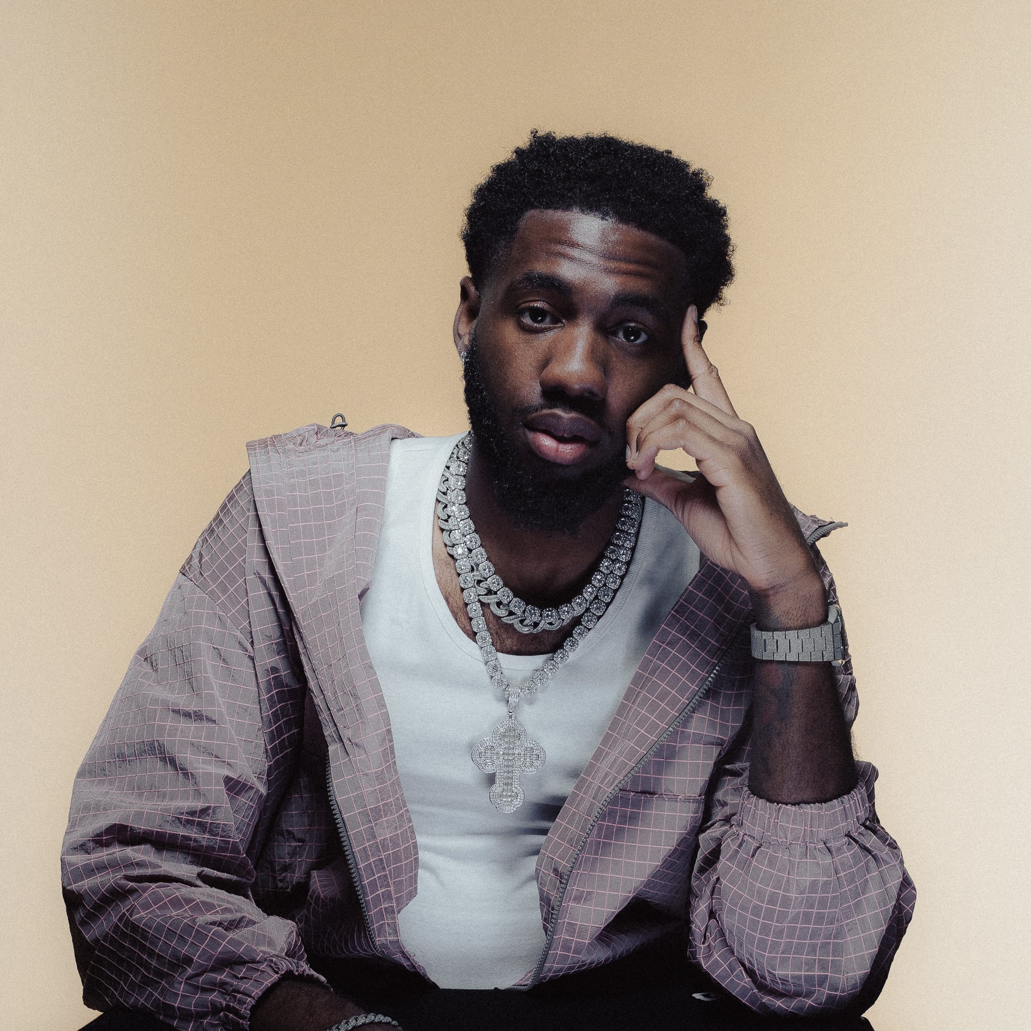 Julio Ade looking directly at the camera, with a finger resting against his temple, on a beige background